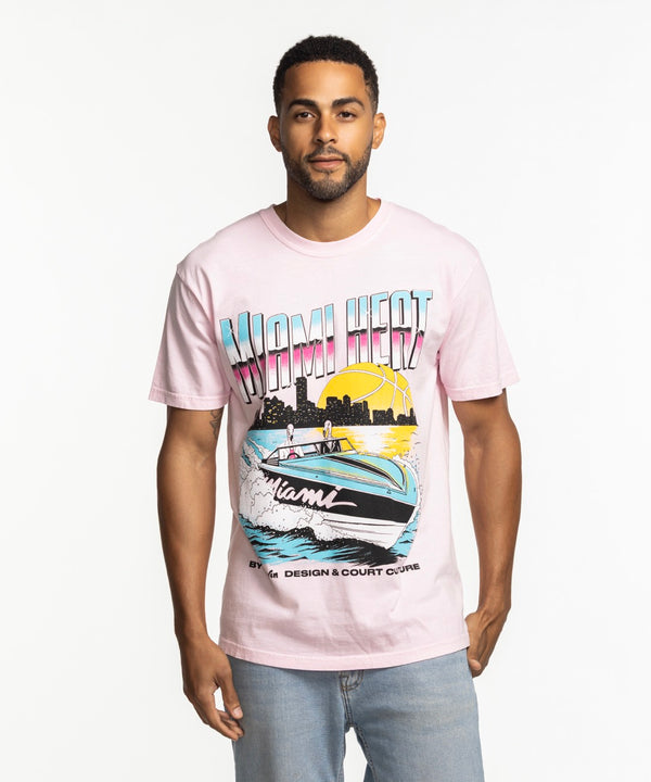 A man stands against a white background wearing the Court Culture x Duvin Boat Tee by Court Culture, a light pink pre-shrunk midweight cotton shirt with a speedboat, sunset, palm trees, and 