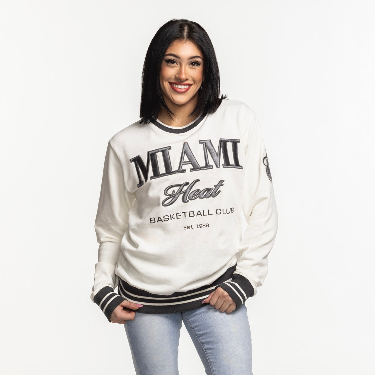 A smiling woman with straight dark hair wears the Sportiqe Miami HEAT Basketball Club Crewneck by Sportiqe and light blue jeans, standing against a plain white background.