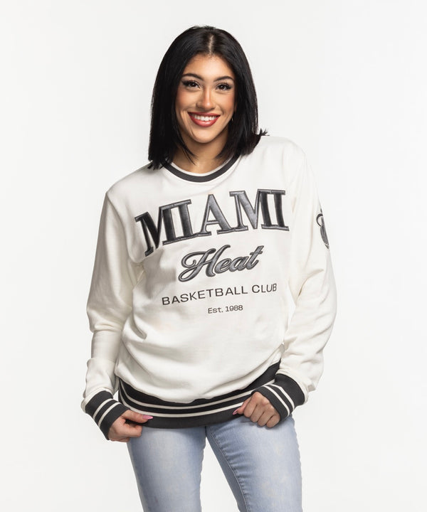 A man wears the Sportiqe Miami HEAT Basketball Club Crewneck, featuring black trim and lettering, styled over light blue jeans against a white background. This crewneck by Sportiqe offers a standard fit for comfort and versatile wear.