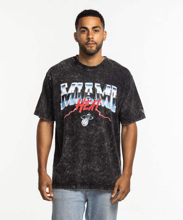A man in a New Era Miami HEAT Original Vice Nights Vintage Tee, featuring black acid-wash 100% cotton and colorful graphic text, stands facing forward against a white background. He has short hair, a trimmed beard, and light blue jeans.