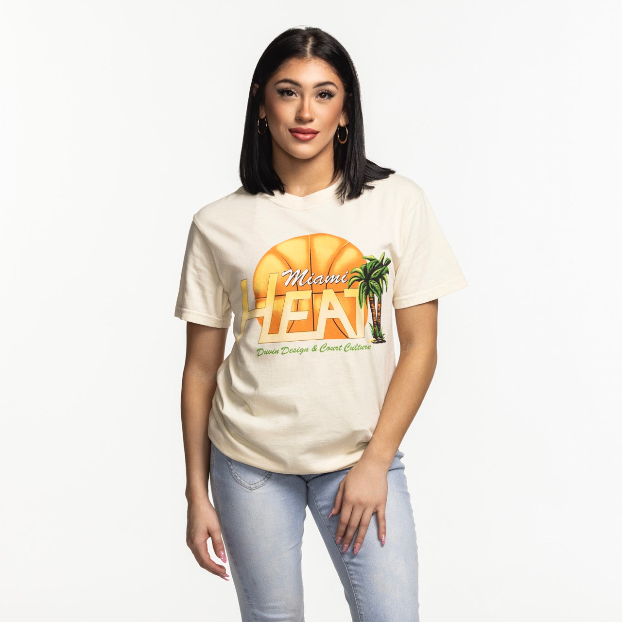 A woman with straight dark hair, hoop earrings, and light blue jeans stands facing forward in a beige Court Culture x Duvin Flames Tee by Court Culture, featuring a basketball and palm tree graphic for stylish comfort.