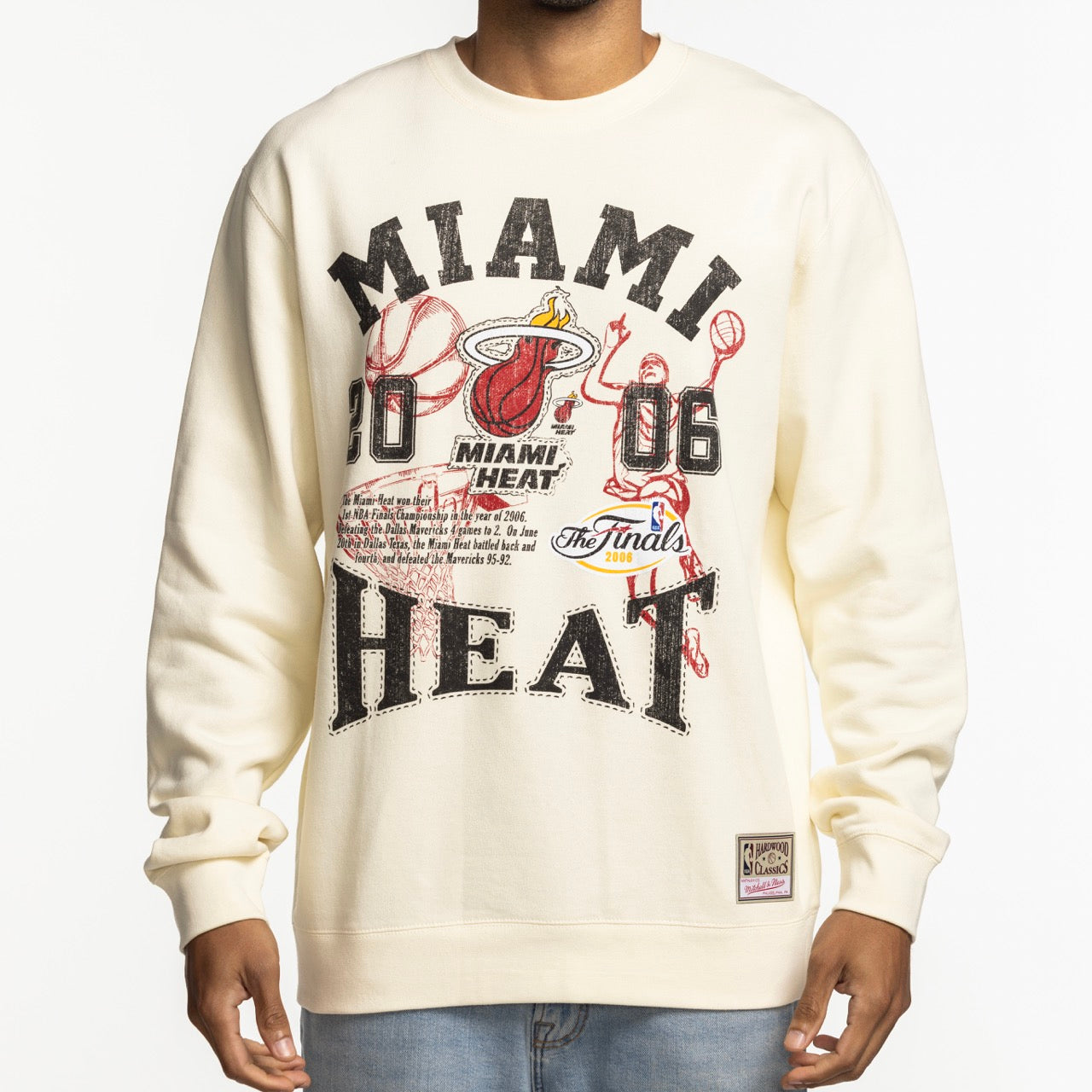 A person wearing the Mitchell & Ness Miami HEAT 2006 Championship Crewneck, cream-colored with bold “MIAMI HEAT,” “2006,” NBA Finals graphics, the Heat logo, basketball elements, and an official NBA patch at the hem. Cotton-polyester; machine washable.