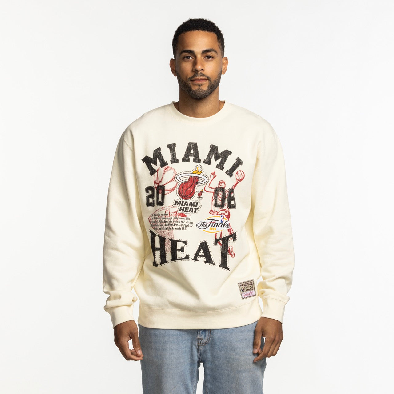 A man faces forward in a Mitchell & Ness Miami HEAT 2006 Championship Crewneck, showcasing team graphics, paired with light blue jeans against a plain white background.