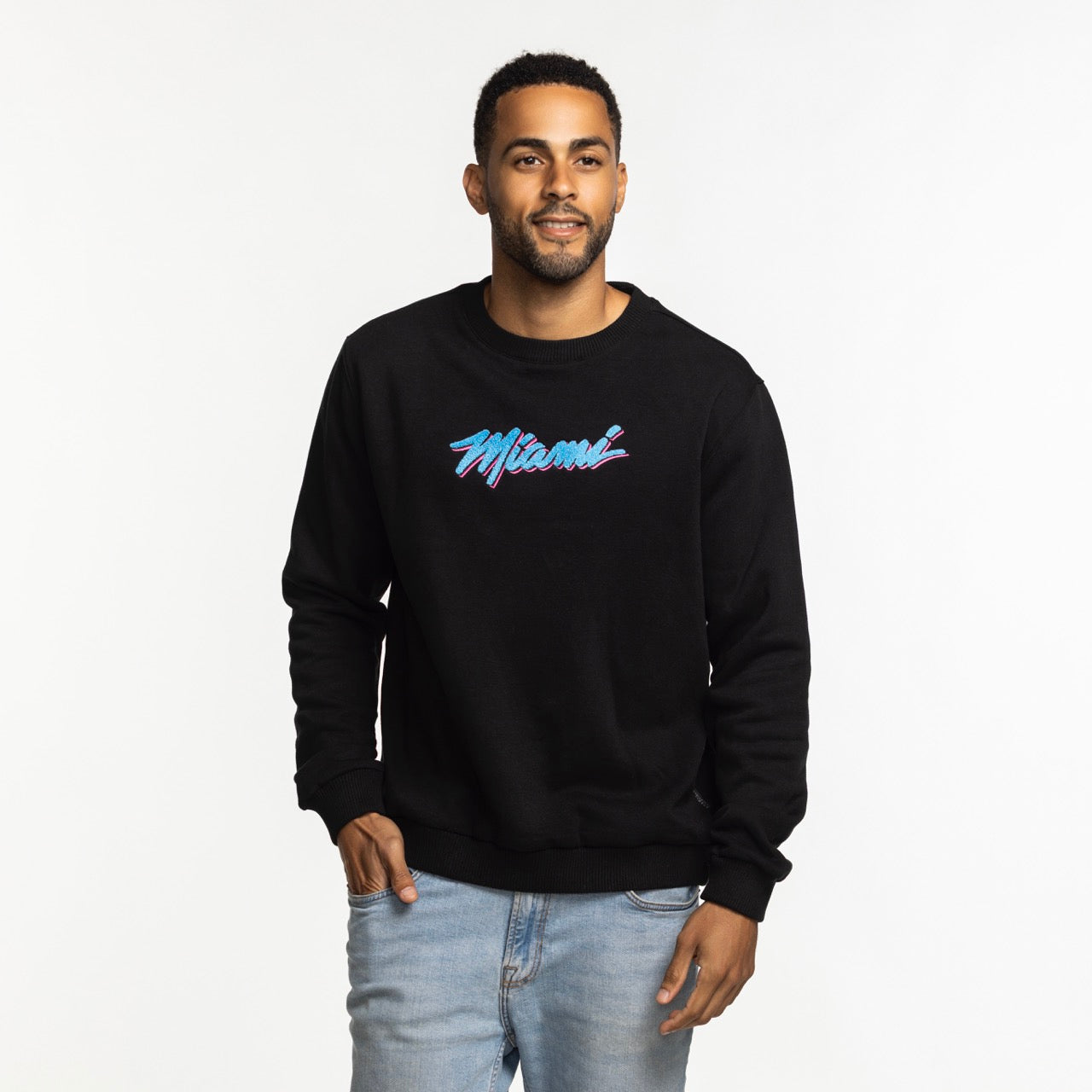 A man smiles with one hand in his pocket, wearing the Stadium Essentials Miami HEAT Original Vice Nights Club Level Crewneck—a black sweatshirt with 