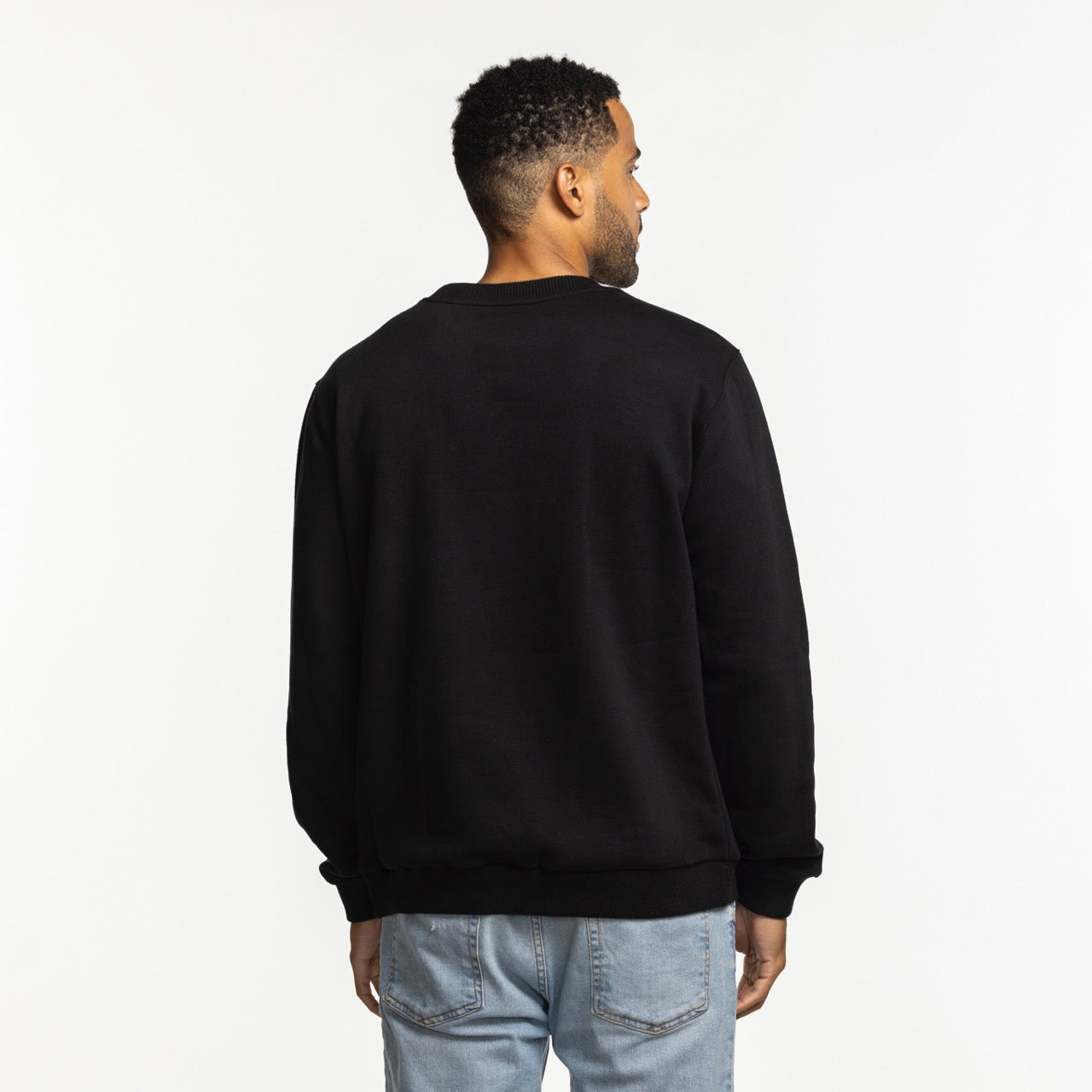 A man faces away from the camera, wearing the Stadium Essentials Miami HEAT Original Vice Nights Club Level Crewneck in black and light blue jeans, standing against a plain white background.