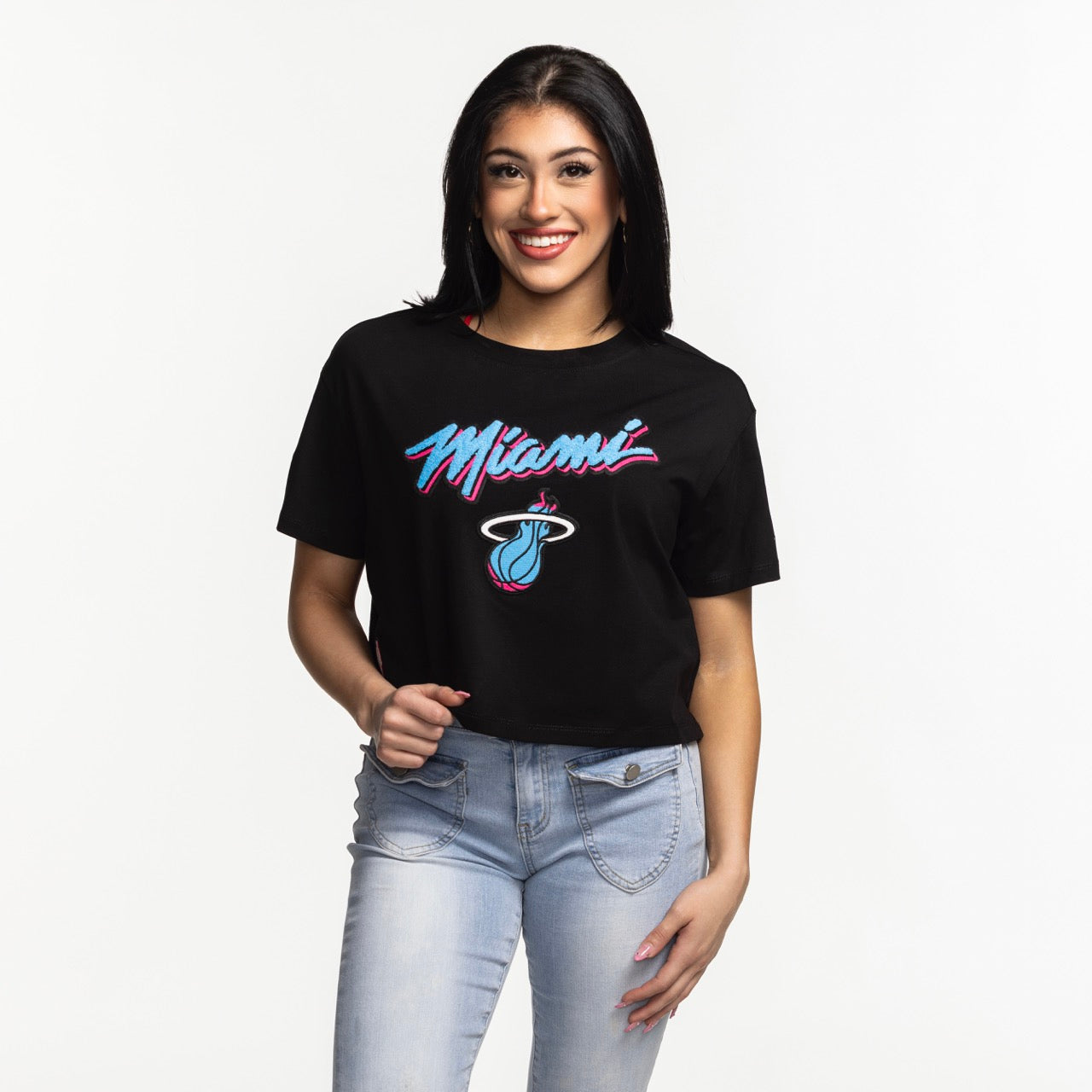 A woman with long dark hair smiles in a Pro Standard Miami HEAT Original Vice Nights Women's Boxy Tee and light blue jeans, standing against a plain white background.