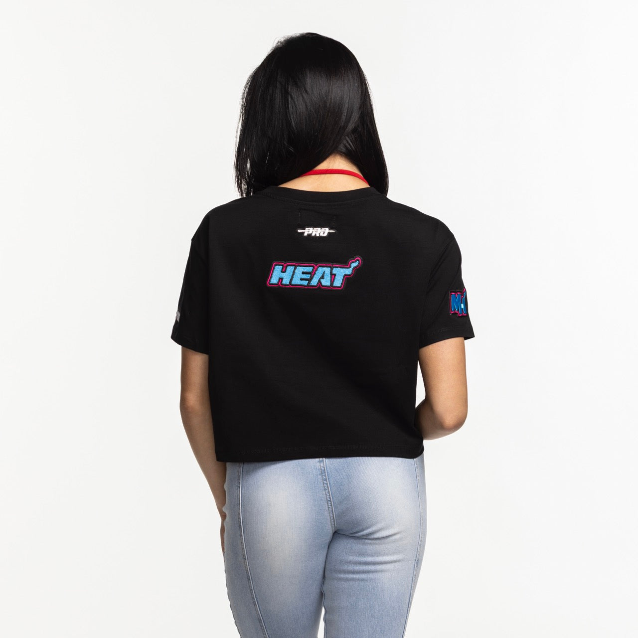 A woman with long dark hair, seen from behind, wears the Pro Standard Miami HEAT Original Vice Nights Women's Boxy Tee and light blue jeans, standing against a white background.