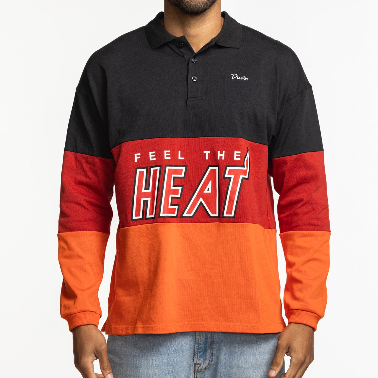 A person wears the Court Culture x Duvin Rugby Polo by Court Culture, a relaxed fit, long-sleeve color-block shirt in black, red, and orange with “FEEL THE HEAT” printed on the front, posed against a plain white background.