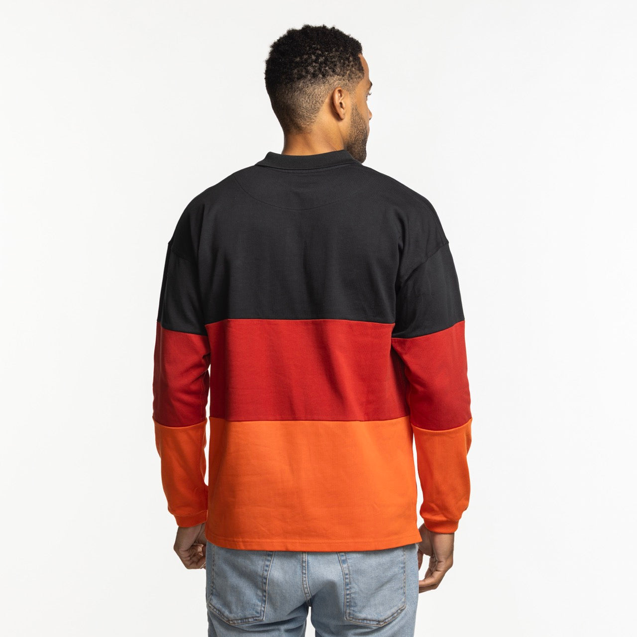 A man faces away from the camera, wearing the Court Culture x Duvin Rugby Polo by Court Culture— a relaxed fit, long-sleeve color-block shirt in black, red, and orange—paired with light blue jeans, set against a plain white background.