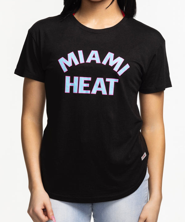 A smiling woman with long dark hair wears the Sportiqe Miami HEAT Original Vice Nights Phoebe Women's Tee by Sportiqe and light blue jeans, standing against a plain white background.