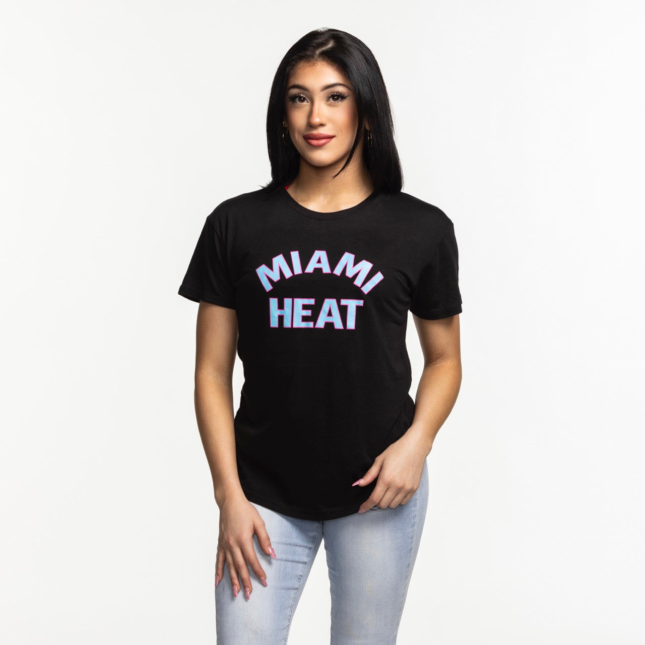 A woman with long dark hair, wearing the Sportiqe Miami HEAT Original Vice Nights Phoebe Women's Tee by Sportiqe and light blue jeans, stands against a plain white background and looks at the camera with a slight smile.