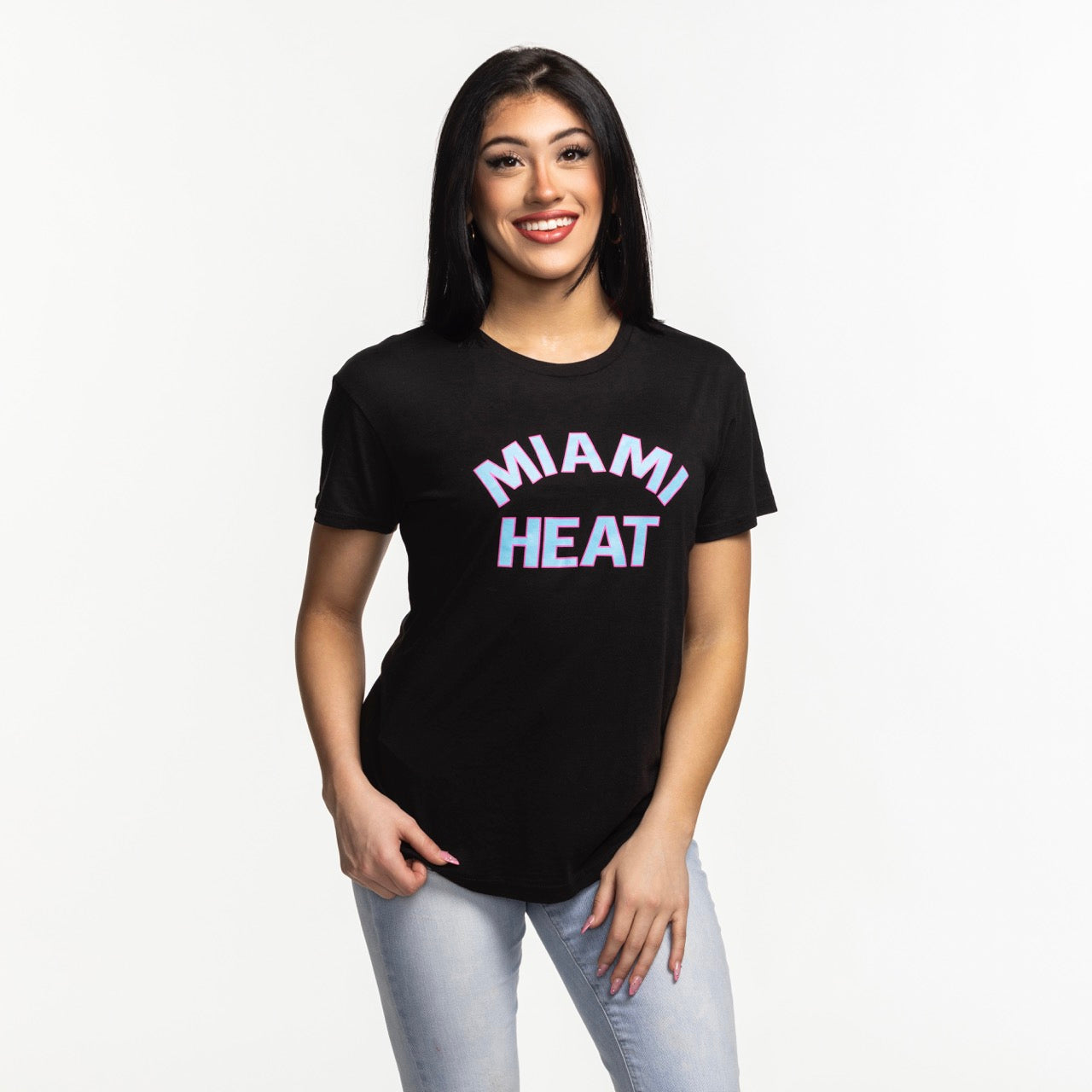 A smiling woman with long dark hair wears the Sportiqe Miami HEAT Original Vice Nights Phoebe Women's Tee by Sportiqe and light blue jeans, standing against a plain white background.