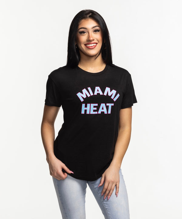 A smiling woman with long dark hair wears the Sportiqe Miami HEAT Original Vice Nights Phoebe Women's Tee by Sportiqe and light blue jeans, standing against a plain white background.