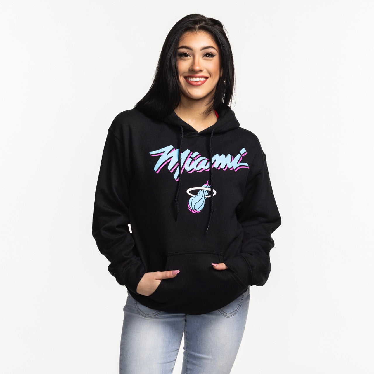 A smiling woman with long dark hair models the Miami HEAT Original Vice Nights Logo Hoodie by Item Of The Game, featuring a pink and blue basketball logo. She pairs it with light blue jeans and stands against a plain white background. Machine wash cold.
