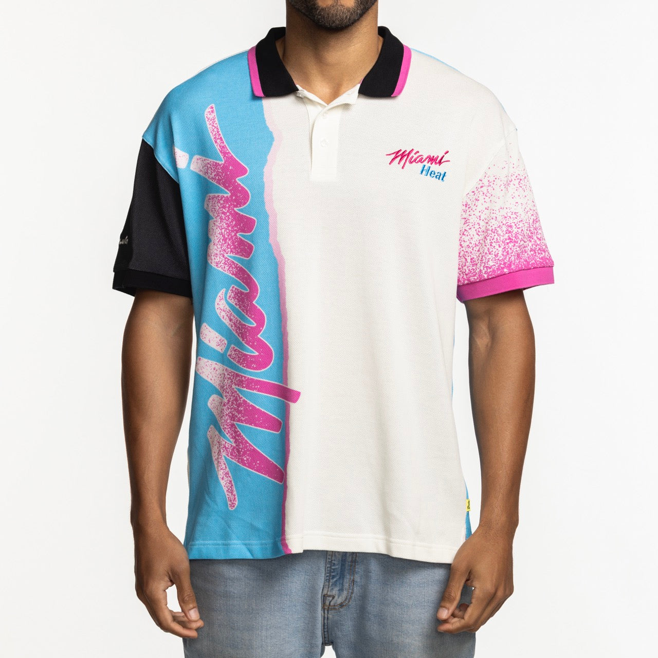 A man wearing the Court Culture x Duvin Sideline Polo by Court Culture, featuring short sleeves, blue and white color-block panels, chest embroidery, large pink Miami script, pink speckles on one sleeve, and Miami Heat text.