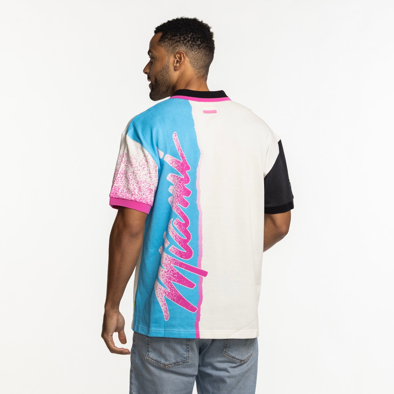 A man with short curly hair and light blue jeans wears the Court Culture x Duvin Sideline Polo—a white short-sleeve cotton shirt with blue/pink accents, chest embroidery, and bold Miami text on the back. Background is plain white.