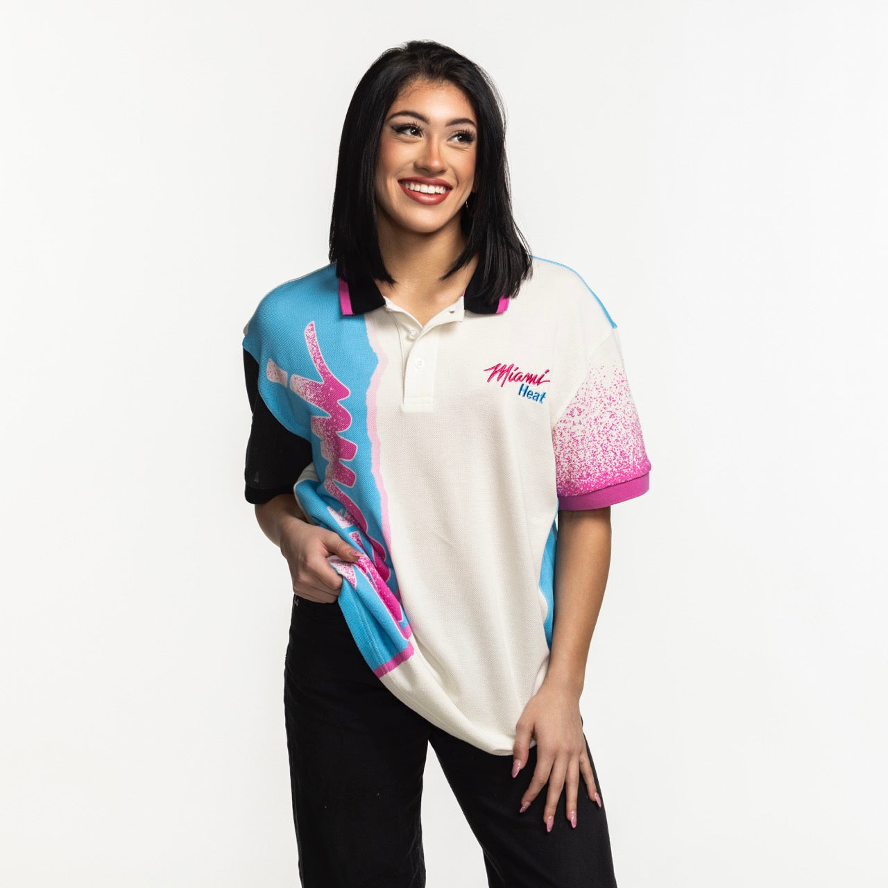 A woman with straight dark hair smiles against a white background, wearing the Court Culture x Duvin Sideline Polo by Court Culture in vibrant colors with chest embroidery, paired with black pants while holding her shirt.