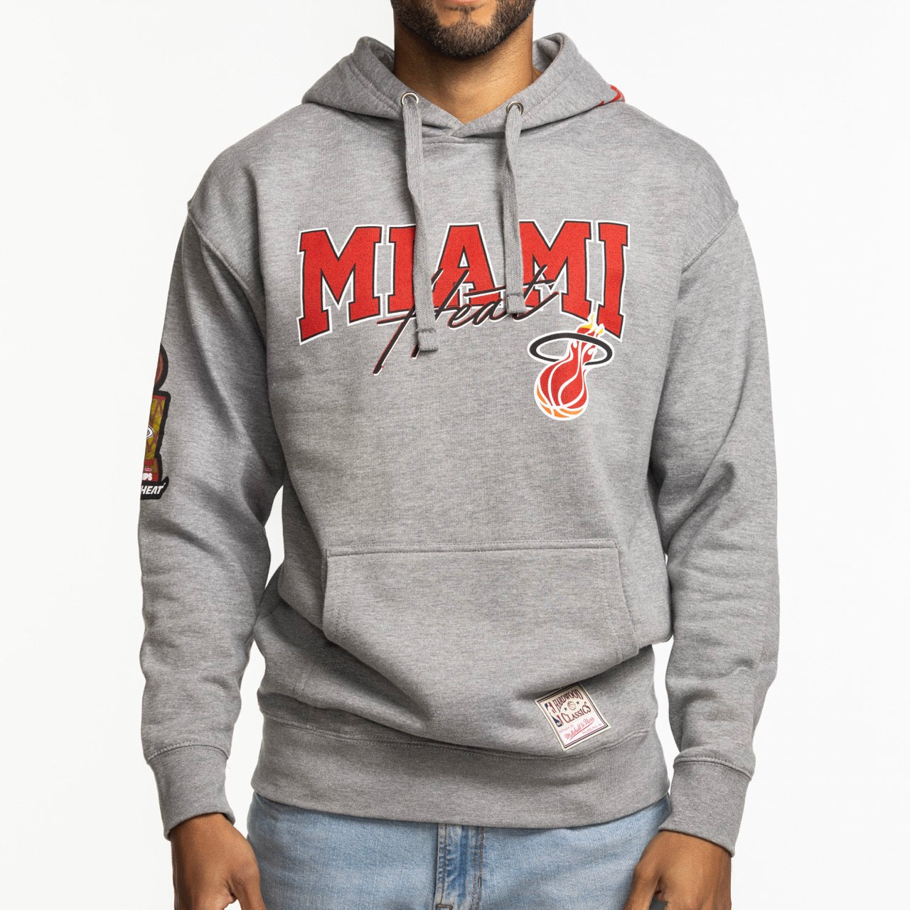 A person wearing the Mitchell & Ness Miami HEAT 2013 Champions Hoodie—a gray hoodie with red MIAMI text, team logo, sleeve patches, front pocket, and drawstrings—made from soft cotton-polyester. Paired with light blue jeans.