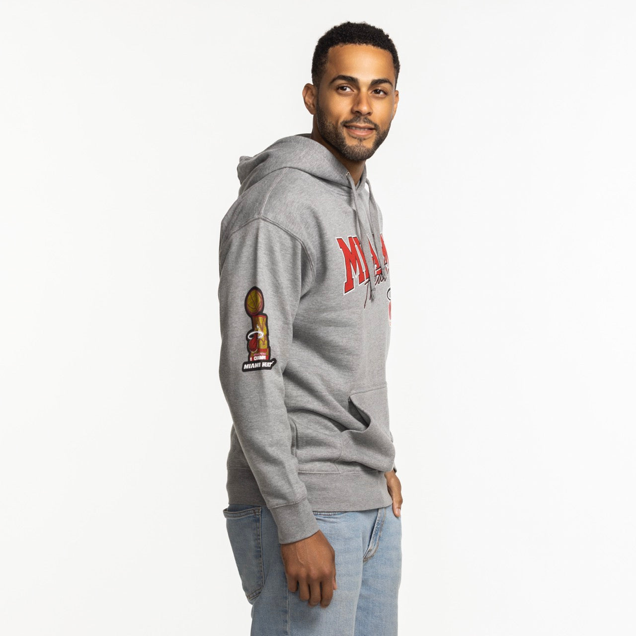A man wears the Mitchell & Ness Miami HEAT 2013 Champions Hoodie, a gray cotton-polyester standard fit with red lettering and a basketball trophy patch on the sleeve, standing sideways and smiling against a white background.