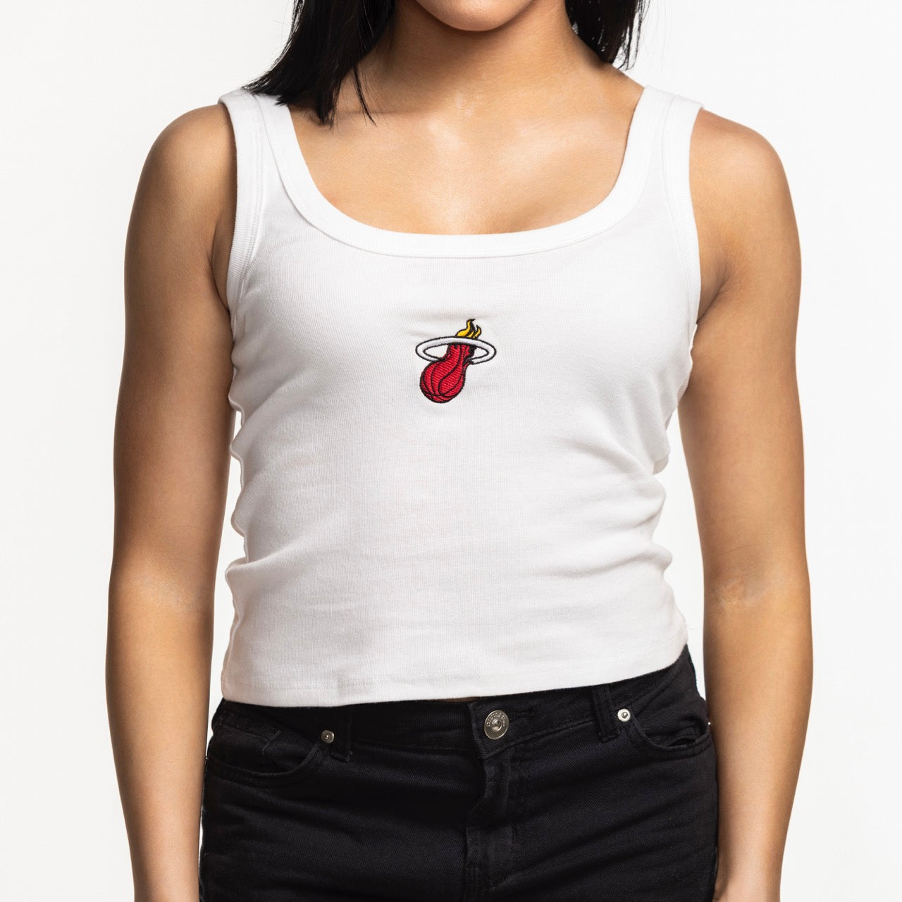A woman wears the Court Culture Embroidered Logo Tank by Court Culture—a standard fit white tank top featuring a Miami Heat basketball logo—paired with black jeans, standing against a plain white background.