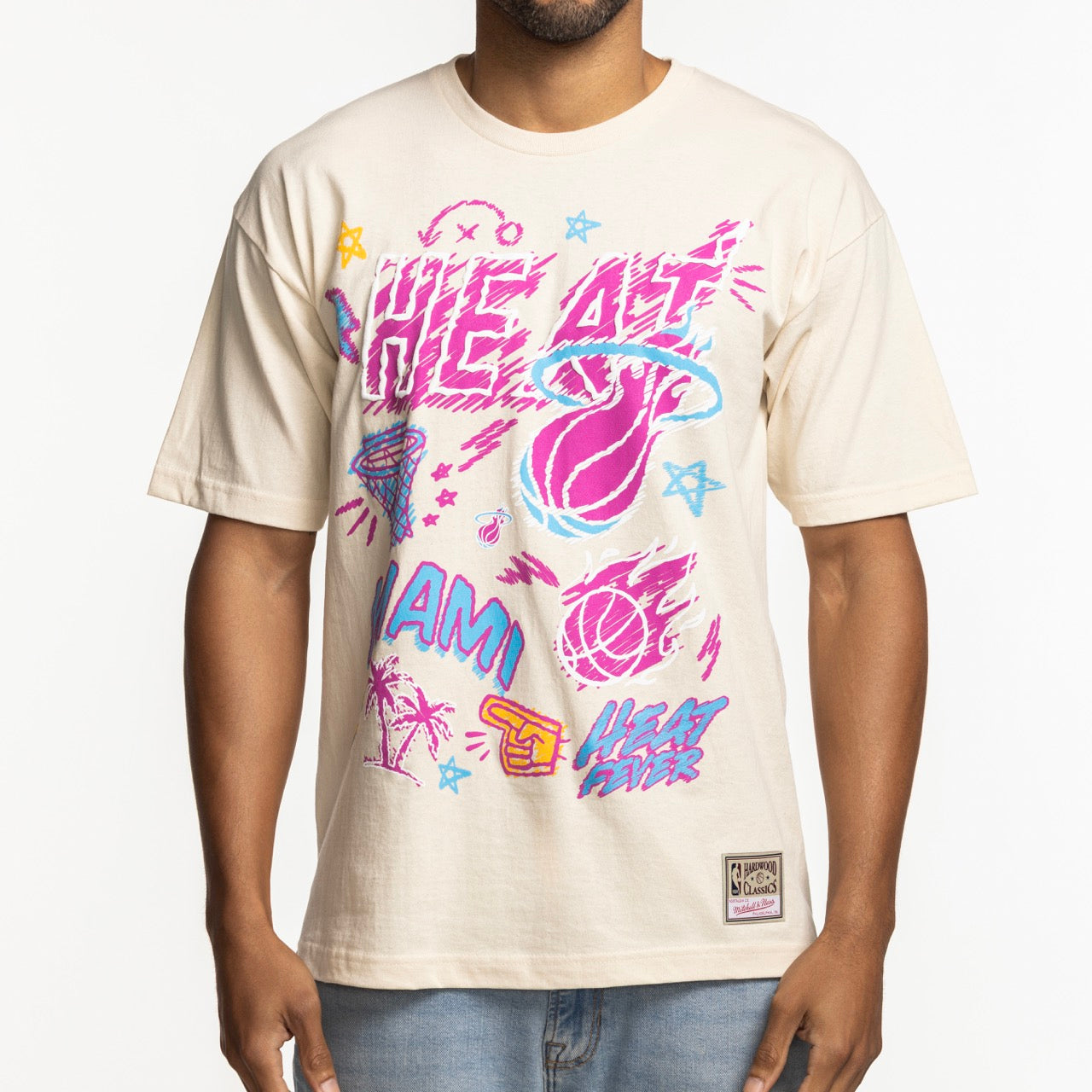 A man wears the Mitchell & Ness Miami HEAT Sketched Out Tee—a standard fit, beige cotton shirt featuring bold pink, blue, and purple retro-style graphics with HEAT logos, palm trees, and basketballs. Machine washable for easy care.
