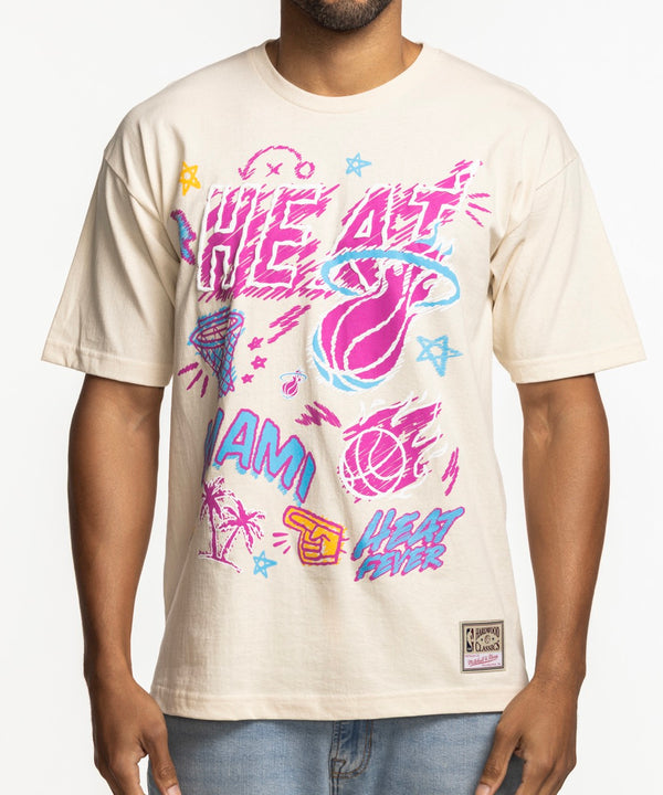 A man wears the Mitchell & Ness Miami HEAT Sketched Out Tee, featuring bold graphics. He pairs it with light blue jeans and stands smiling against a white background. This cotton tee by Mitchell & Ness is machine washable for easy care.