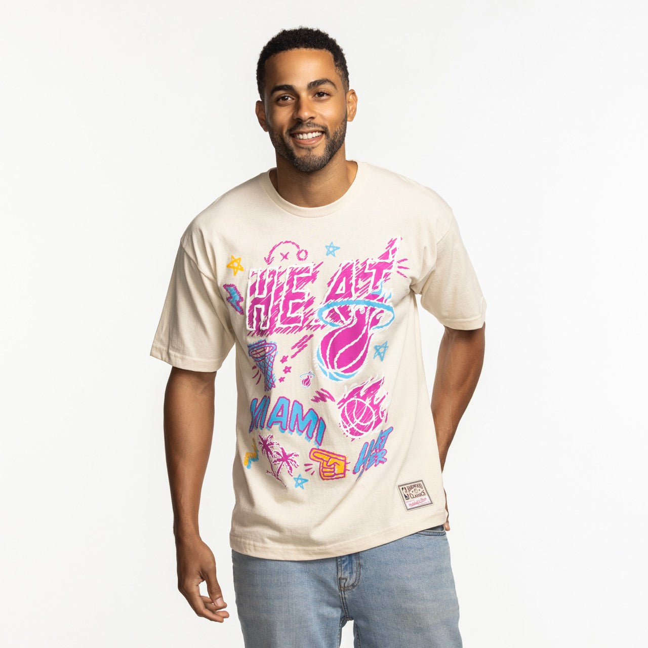A man wears the Mitchell & Ness Miami HEAT Sketched Out Tee, featuring bold graphics. He pairs it with light blue jeans and stands smiling against a white background. This cotton tee by Mitchell & Ness is machine washable for easy care.