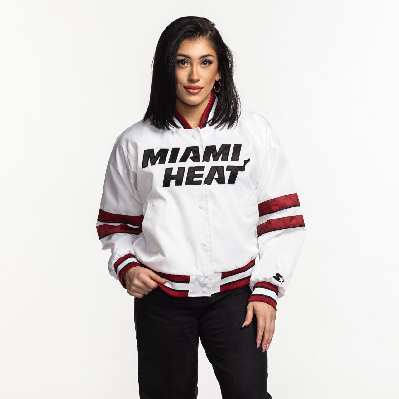 A woman with straight black hair stands against a white background wearing the G-III Sports Miami HEAT Interception Women's Jacket in white with red accents, black pants, and gold hoop earrings. She faces forward with her hands in the jacket pockets.