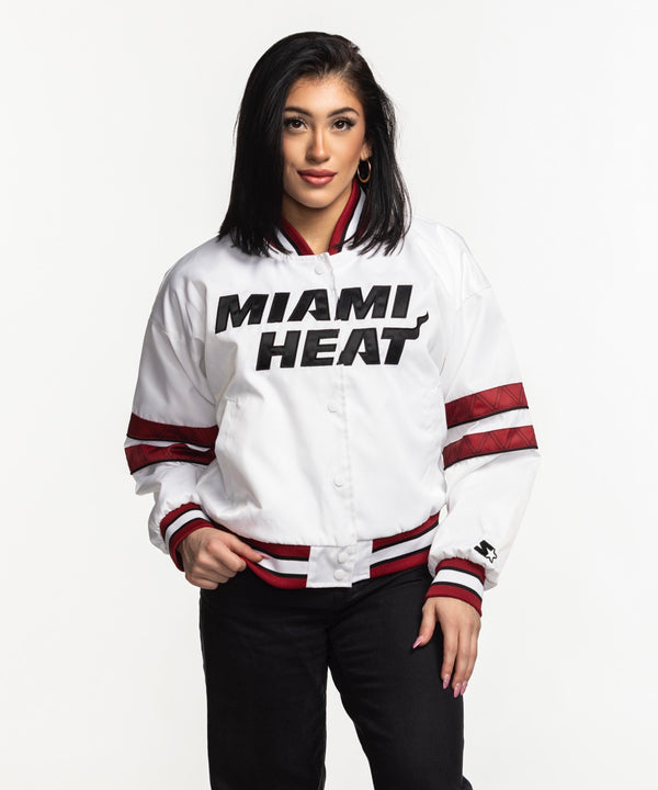 A woman with straight black hair stands against a white background wearing the G-III Sports Miami HEAT Interception Women's Jacket in white with red accents, black pants, and gold hoop earrings. She faces forward with her hands in the jacket pockets.