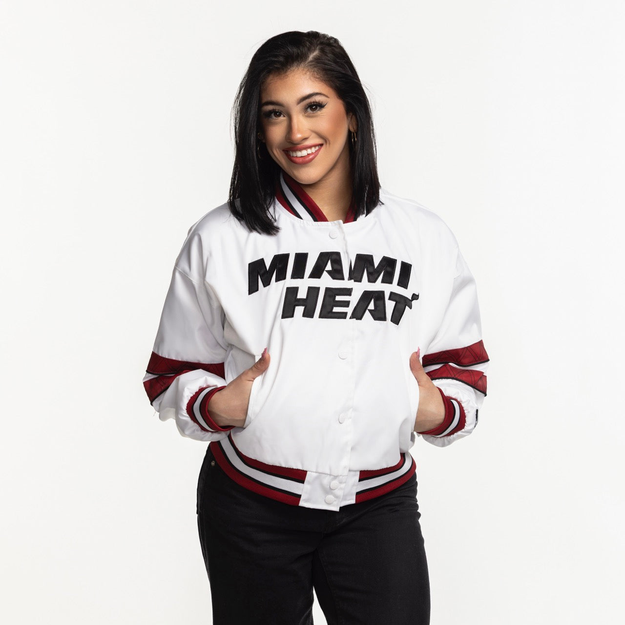 A smiling woman with dark hair wears the G-III Sports Miami HEAT Interception Women's Jacket, made from durable polyester with red and black accents, standing with her hands in the pockets against a white background. Machine washable.