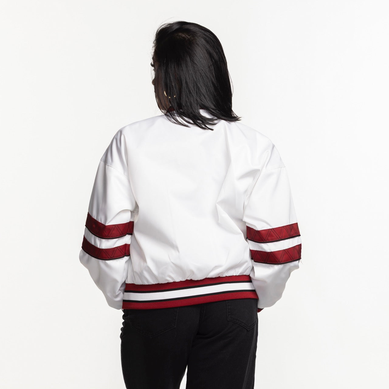 A person faces away, wearing a G-III Sports Miami HEAT Interception Women's Jacket—standard fit, white polyester with red stripes on the sleeves and waistband—with black pants. The jacket is machine washable. Plain white background.