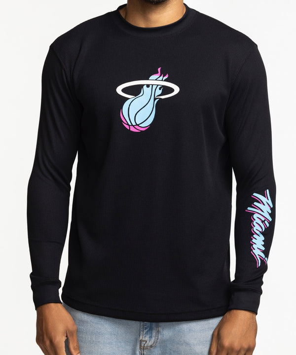 A man wears the Stadium Essentials Miami HEAT Original Vice Nights Long Sleeve Tee, a black polyester-spandex shirt with colorful team graphics, paired with light blue jeans. Machine wash cold for best results.