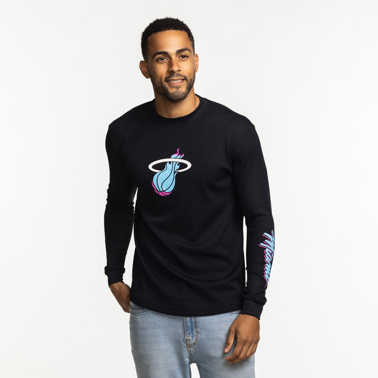 A man wears the Stadium Essentials Miami HEAT Original Vice Nights Long Sleeve Tee, a black polyester-spandex shirt with colorful team graphics, paired with light blue jeans. Machine wash cold for best results.