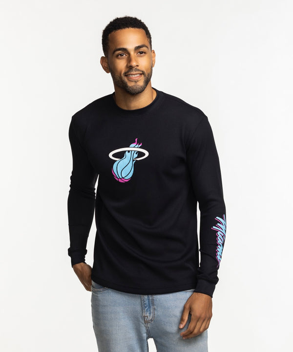 A man wears the Stadium Essentials Miami HEAT Original Vice Nights Long Sleeve Tee, a black polyester-spandex shirt with colorful team graphics, paired with light blue jeans. Machine wash cold for best results.