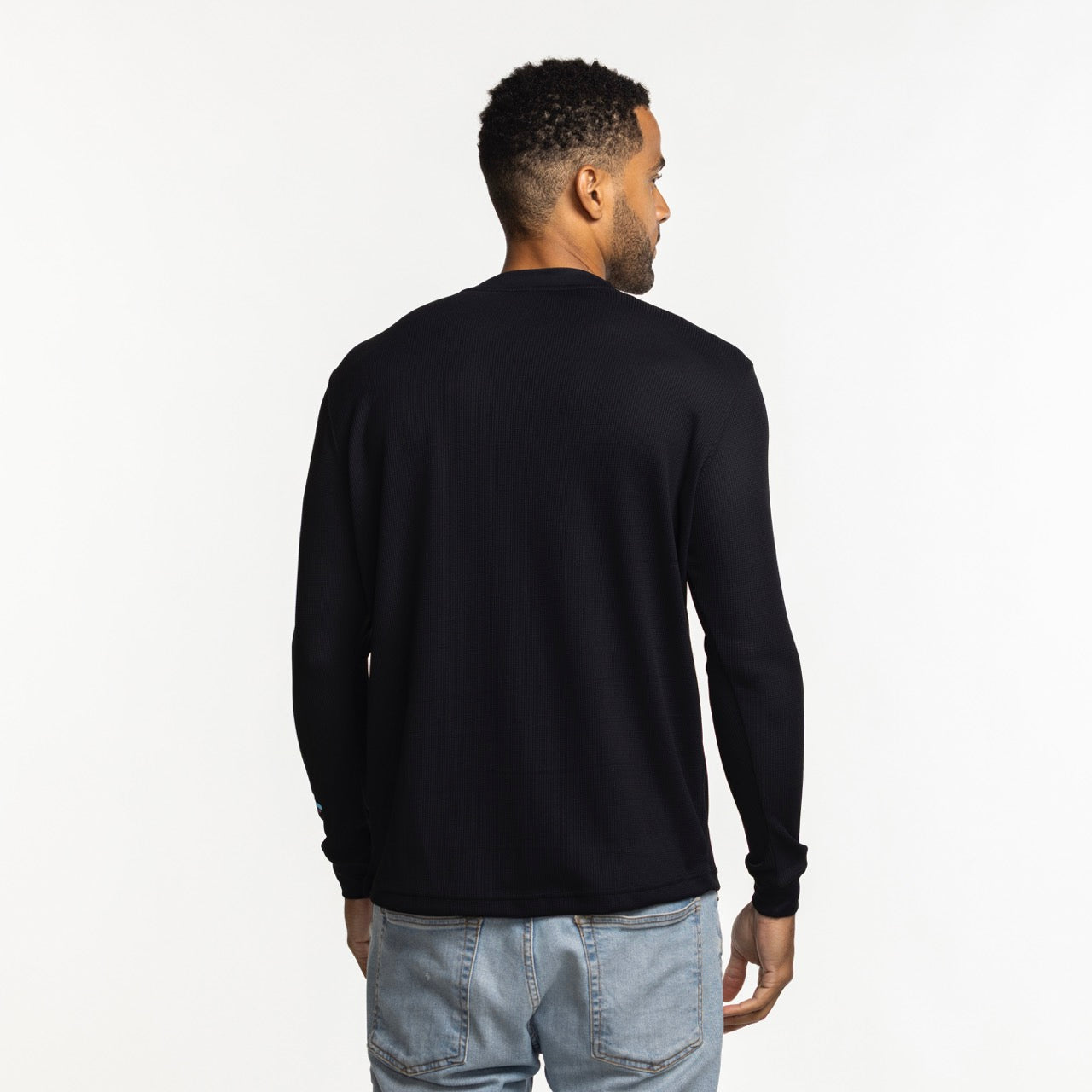 A man with short curly hair is standing with his back to the camera, wearing a Stadium Essentials Miami HEAT Original Vice Nights Long Sleeve Tee and light blue jeans, against a plain white background.