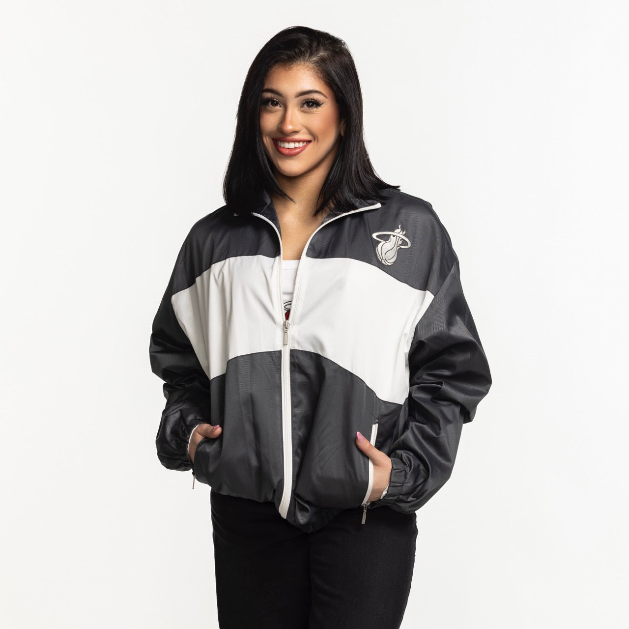 A woman with straight dark hair smiles at the camera, wearing a black and white Sportiqe Miami HEAT Basketball Club Jacket with her hands in the pockets, standing against a plain white background.