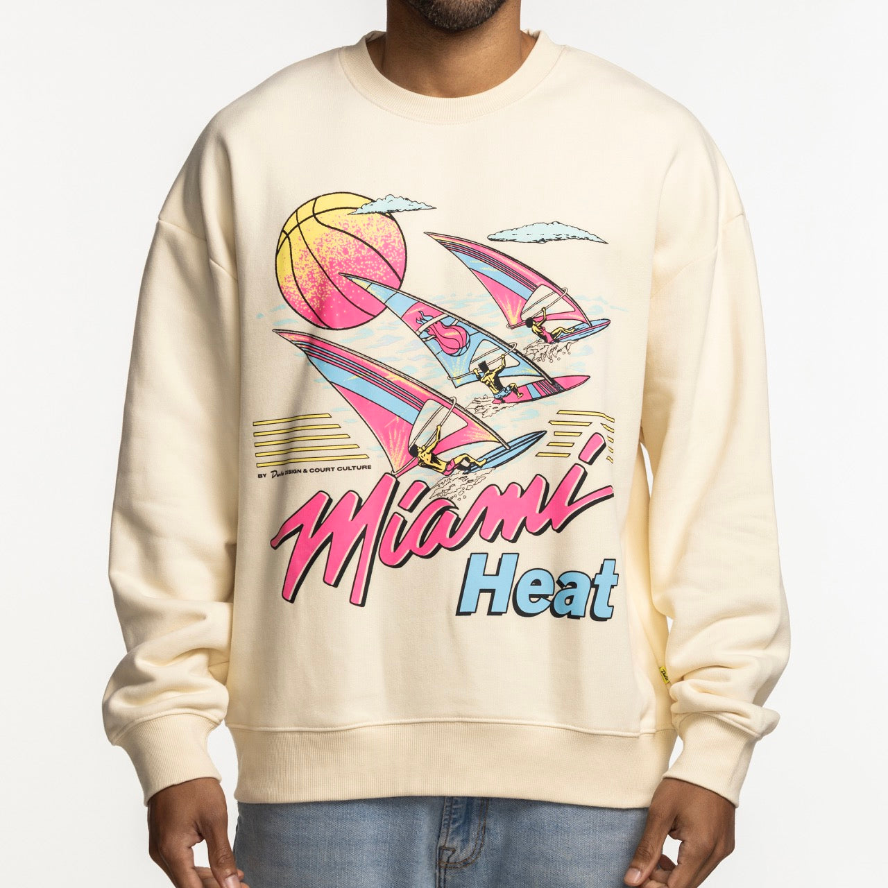 A person wears the Court Culture x Duvin Ivory Sweater—a cream heavyweight fleece with a retro Miami Heat graphic featuring sailboats, a basketball sun, and pink and blue accents. Only their torso is visible. Brand: Court Culture.