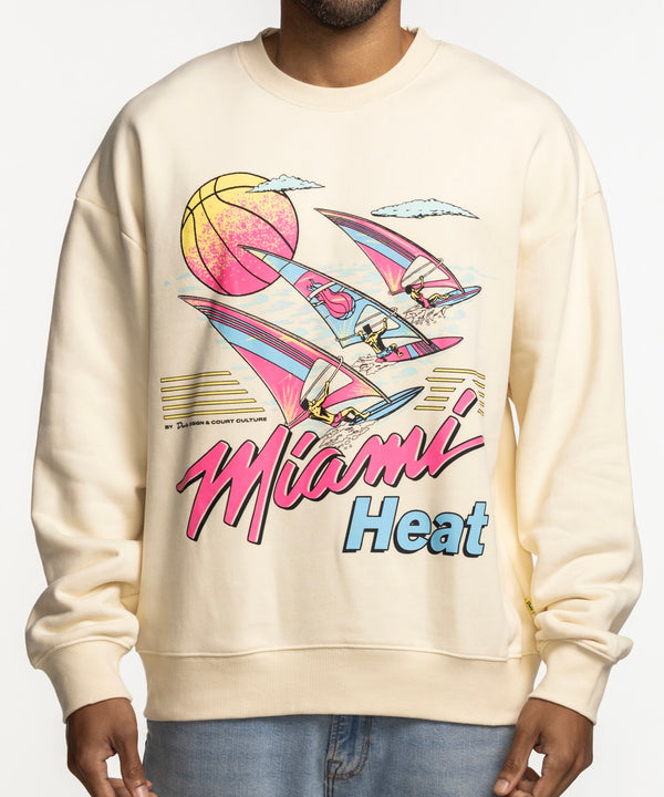 A man smiles against a white background wearing the Court Culture x Duvin Ivory Sweater by Court Culture, a relaxed fit crewneck in cream with a colorful retro Miami Heat graphic featuring a basketball, flamingos, and palm trees.