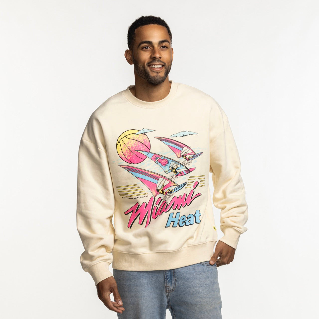 A man smiles against a white background wearing the Court Culture x Duvin Ivory Sweater by Court Culture, a relaxed fit crewneck in cream with a colorful retro Miami Heat graphic featuring a basketball, flamingos, and palm trees.