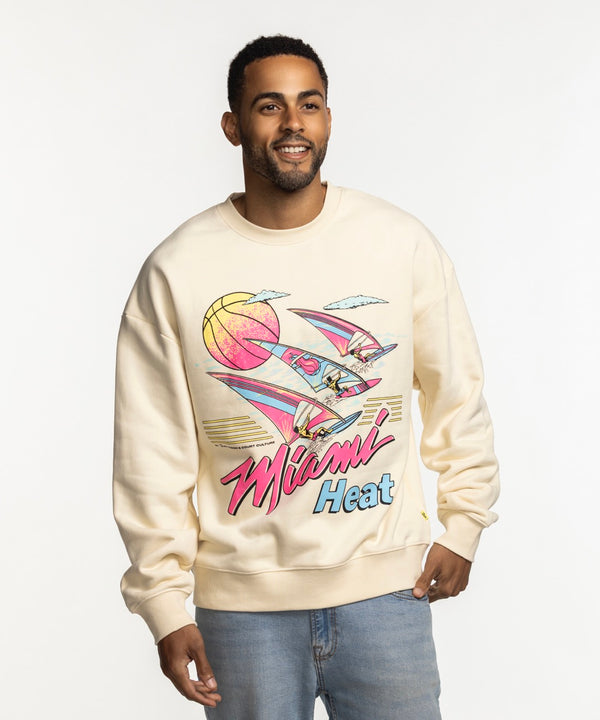 A man smiles against a white background wearing the Court Culture x Duvin Ivory Sweater by Court Culture, a relaxed fit crewneck in cream with a colorful retro Miami Heat graphic featuring a basketball, flamingos, and palm trees.