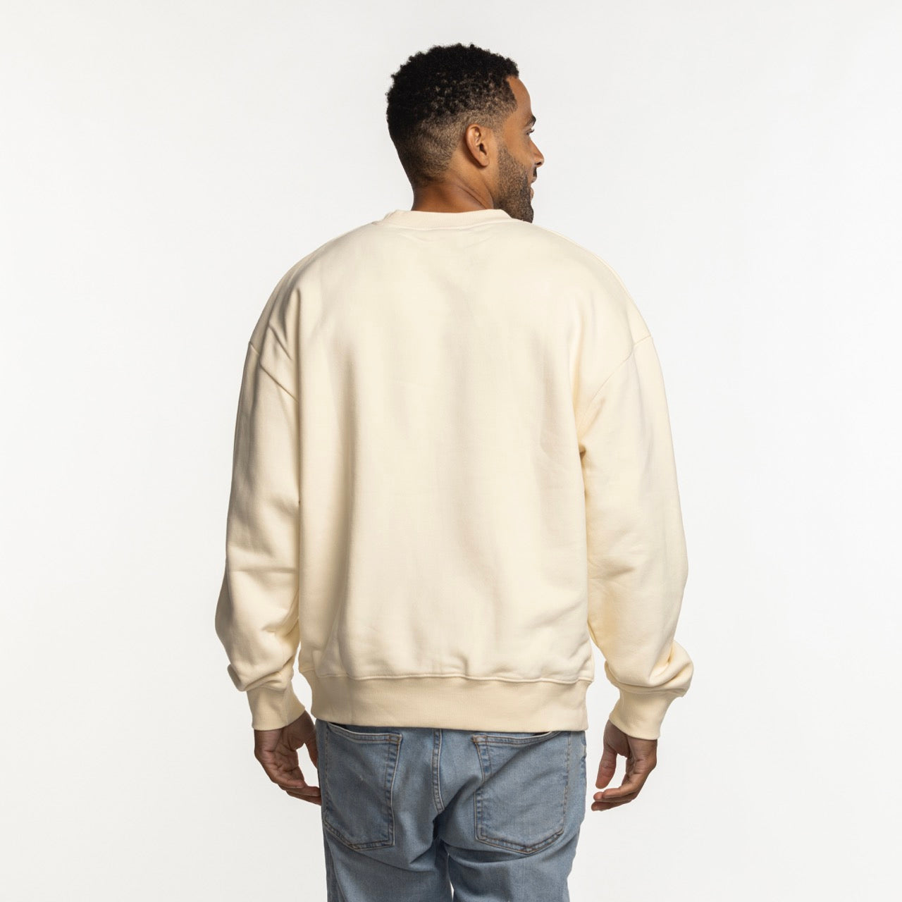 A man wears the Court Culture x Duvin Ivory Sweater by Court Culture with light blue jeans, standing with his back to the camera against a plain white background.