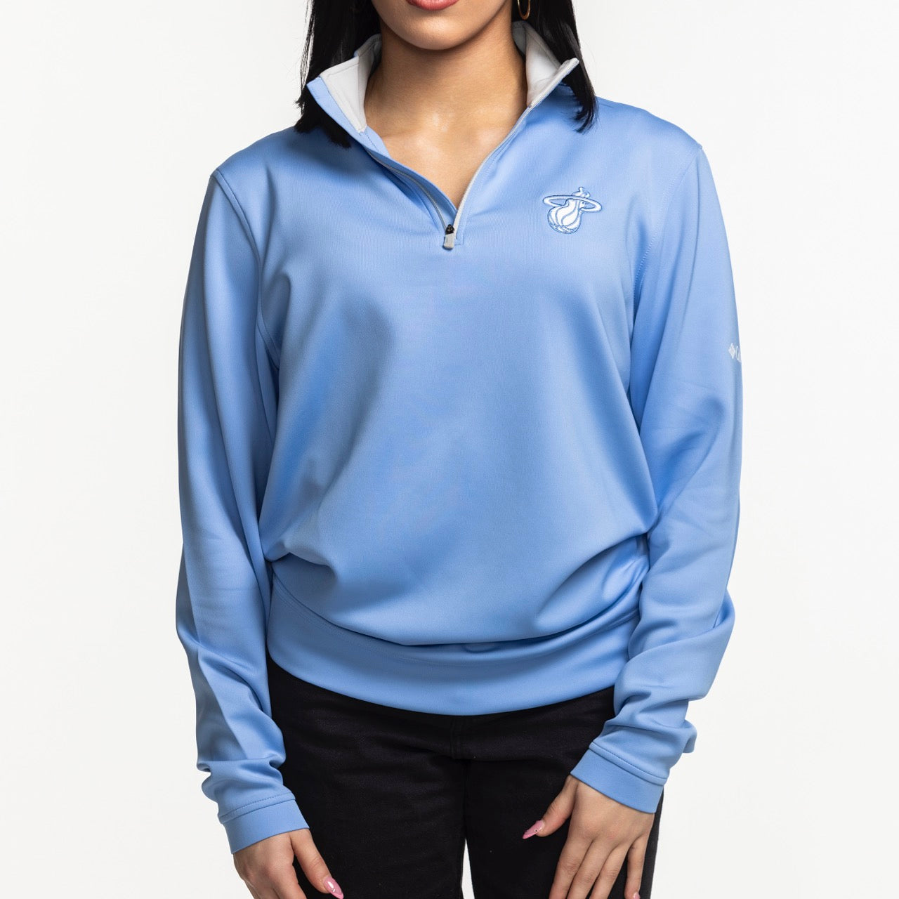 A person wears the Columbia Miami HEAT Original Vice Nights Women's 1/4 Zip by Columbia Outdoor—a light blue, relaxed fit quarter-zip with the Miami Heat logo—paired with black pants, hands in front against a plain white background.