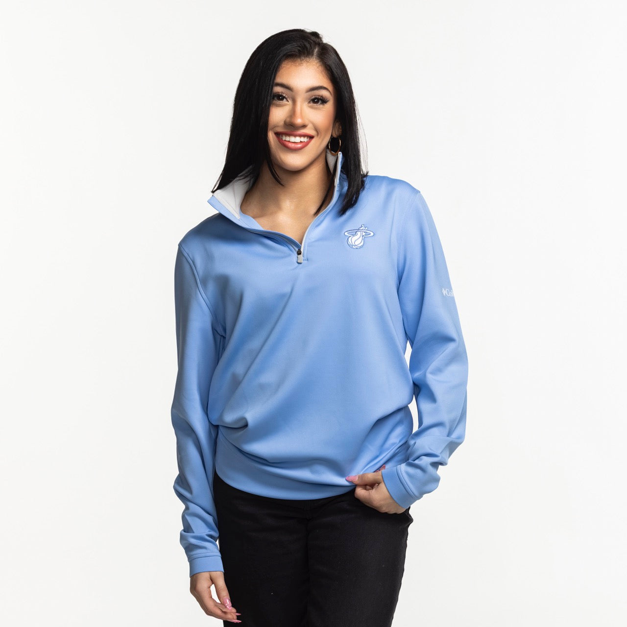 A woman with straight dark hair smiles while wearing the Columbia Outdoor Miami HEAT Original Vice Nights Women's 1/4 Zip pullover in light blue and black pants, standing against a plain white background.