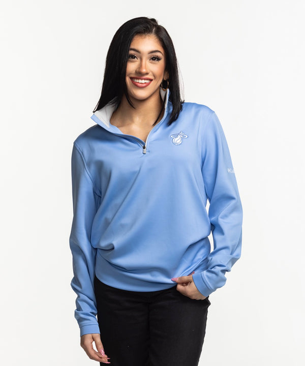 A woman with straight dark hair smiles while wearing the Columbia Outdoor Miami HEAT Original Vice Nights Women's 1/4 Zip pullover in light blue and black pants, standing against a plain white background.