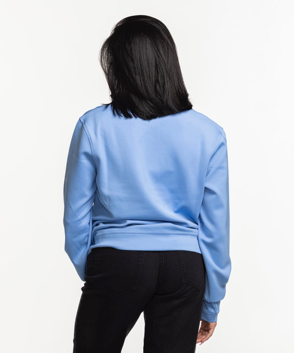 A woman with straight dark hair smiles while wearing the Columbia Outdoor Miami HEAT Original Vice Nights Women's 1/4 Zip pullover in light blue and black pants, standing against a plain white background.