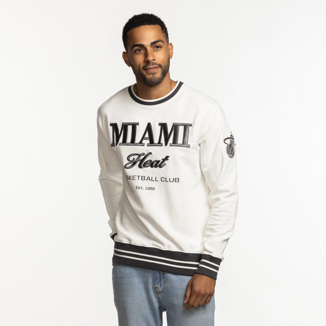 A man wears the Sportiqe Miami HEAT Basketball Club Crewneck, featuring black trim and lettering, styled over light blue jeans against a white background. This crewneck by Sportiqe offers a standard fit for comfort and versatile wear.