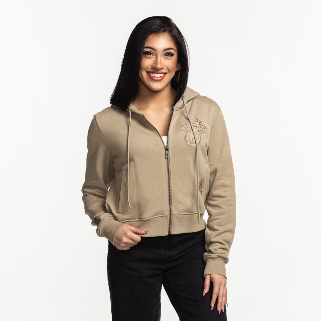 A woman with long dark hair smiles while wearing the Pro Standard Miami HEAT Original Vice Nights Beige Women's Hoodie over a white top and black pants, standing against a plain white background.