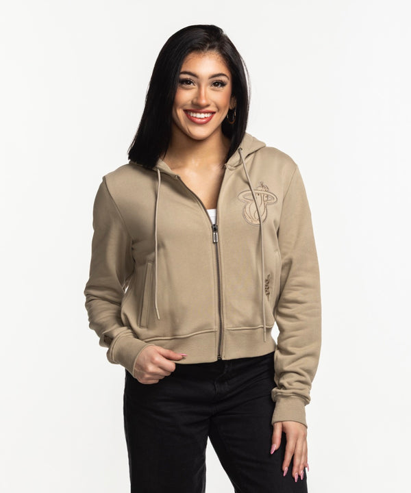A woman with long dark hair smiles while wearing the Pro Standard Miami HEAT Original Vice Nights Beige Women's Hoodie over a white top and black pants, standing against a plain white background.