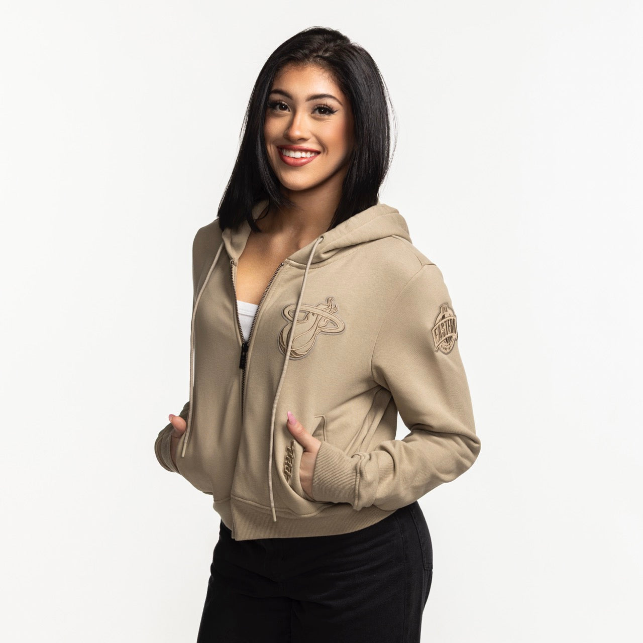A young woman with straight dark hair smiles at the camera, wearing a Pro Standard Miami HEAT Original Vice Nights Beige Women's Hoodie over a white top and black pants, against a plain white background.