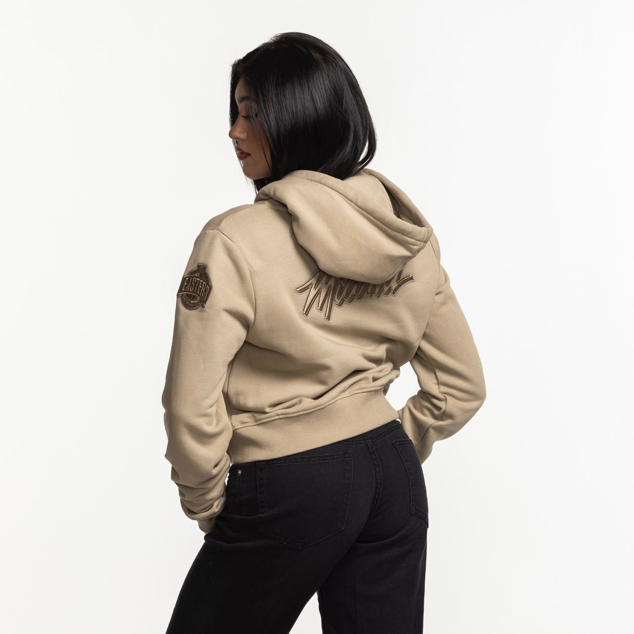A woman with straight black hair models the Pro Standard Miami HEAT Original Vice Nights Beige Women's Hoodie, featuring embroidered back text and a sleeve patch, black jeans, and a cropped fit against a white background.